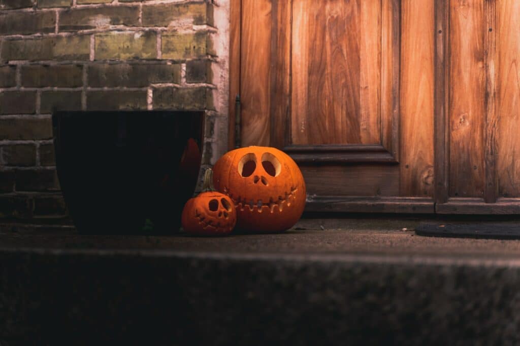 Two jack'o'lanterns on a doorstep next to a treat bucket.