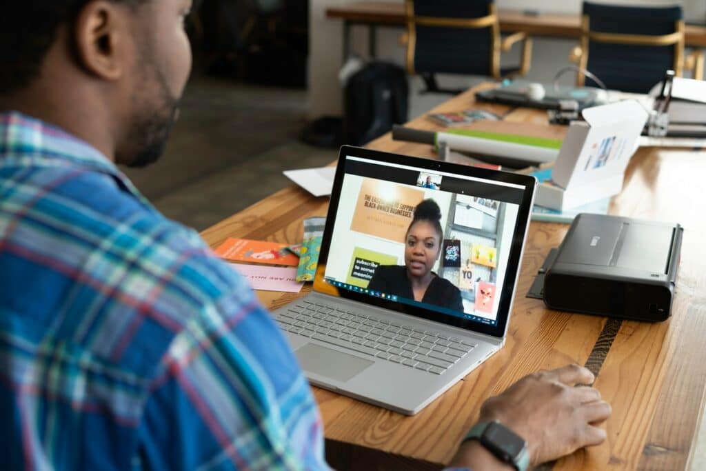 A person, viewed from behind, is on a video conferencing app talking to another person on a laptop that is on their kitchen table. 
