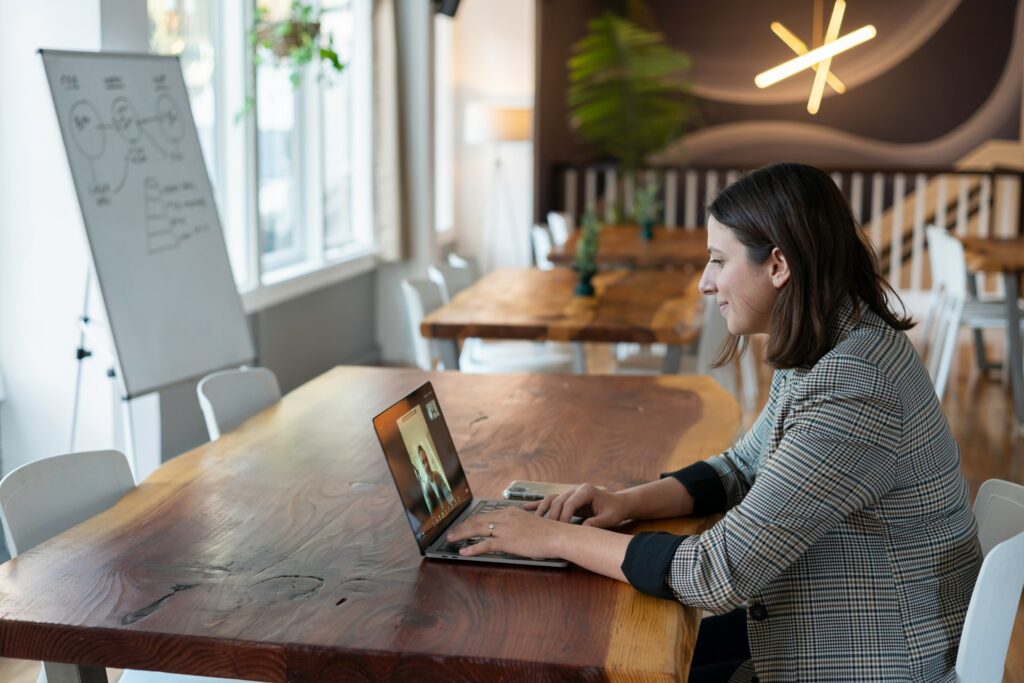 A person is sat in a cafe at a desk with a whiteboard in front of them, looking at a video conference app on their laptop, which is on a desk with someone on a call with them.