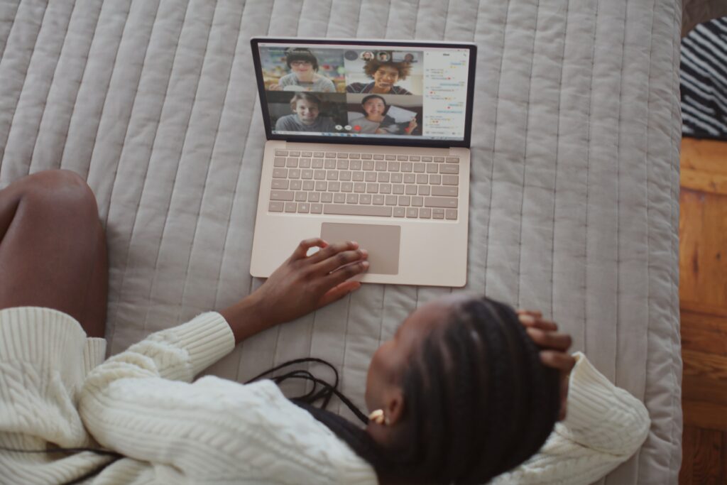 A person lays on a bed looking at their laptop which is displaying a web conference app with multiple people displayed on the screen.