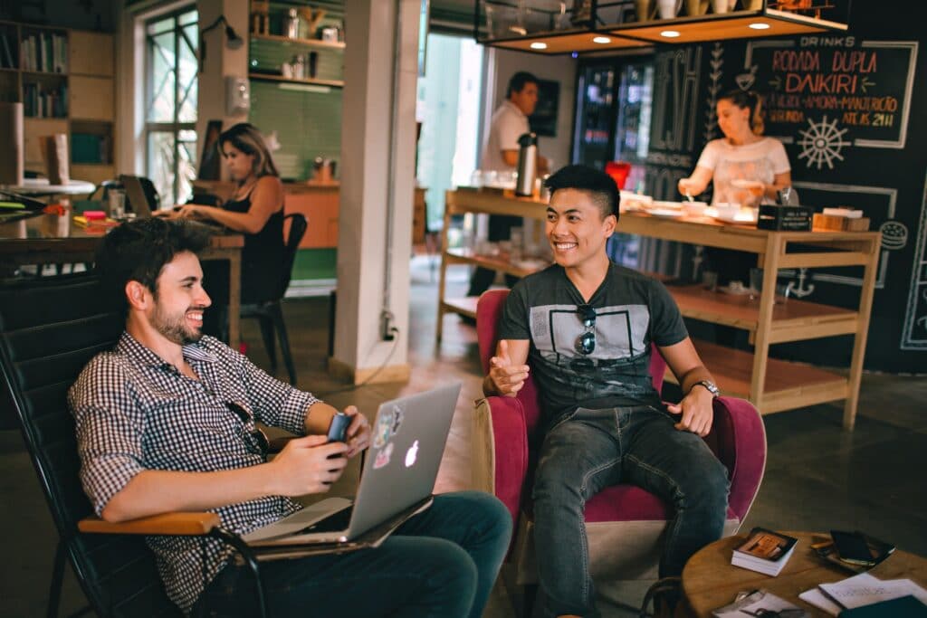 Two people sat in a hybrid meeting space with people behind them as they talk.