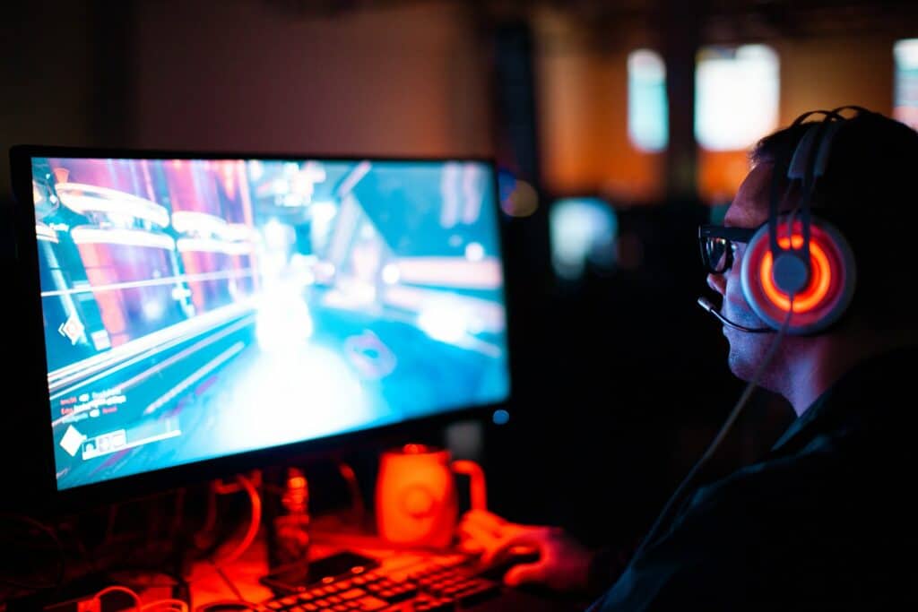 A person with an RGB gaming headset on, streaming a game on their monitor looking to stream with guests with a keyboard and Star Wars mug on their desk.