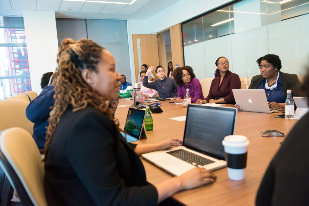 Multiple people sat around a large meeting room table with their laptops open, looking towards the far end of the table where someone is pitching a non-profit.