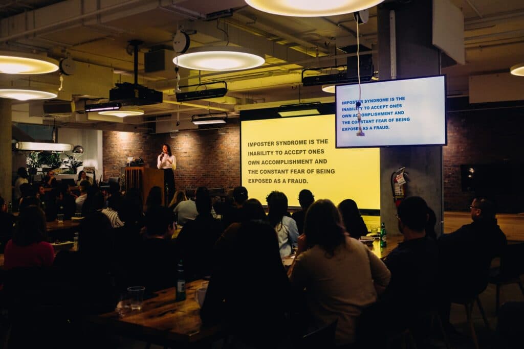 A yellow slide with black text being show to a large room of people. Even for simple slides, like this yellow background, makes a big difference for how people feel viewing it for color theory in presentations!
