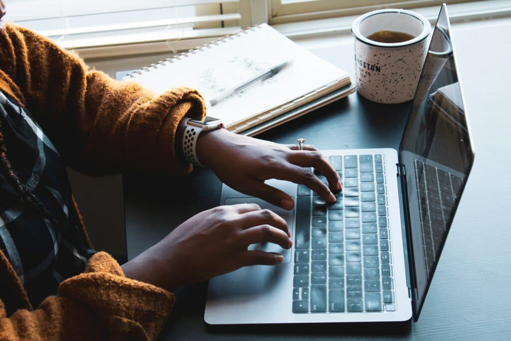 A person sat at a desk, using their laptop with notes and a mug of tea on the desk as they learn how to write video tutorials