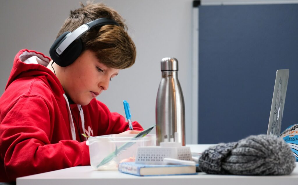 A child sat at their desk wearing headphones with a laptop in front of them while they write in a school book, on their desk is a water bottle, pens, a hat, another note book and some snacks looking for ways to improve teaching.