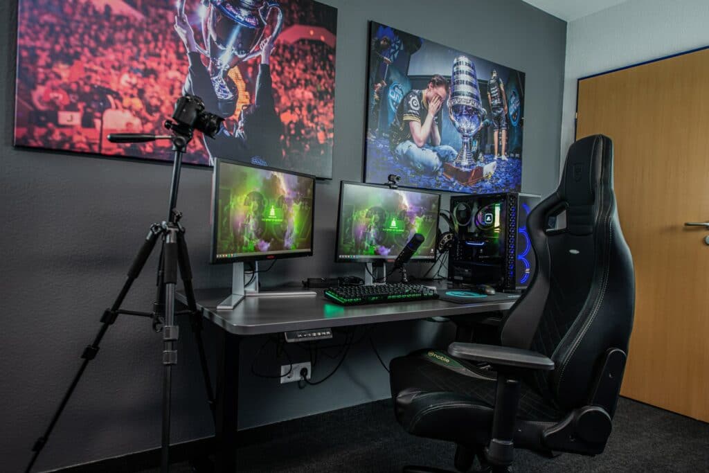 A large gaming chair is sat at a desk with two monitors, a PC, a microphone, a webcam. On the wall behind it are pictures of esports players winning cups. A DSLR camera is on a tripod next to the desk. The desk, is a standing desk, don't forget to stretch when you're looking to improve your viewership!