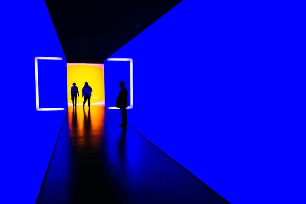 Live stream art like this large physical installation of lights illuminating a hall with two people entering from one end bathed in orange and blue light.