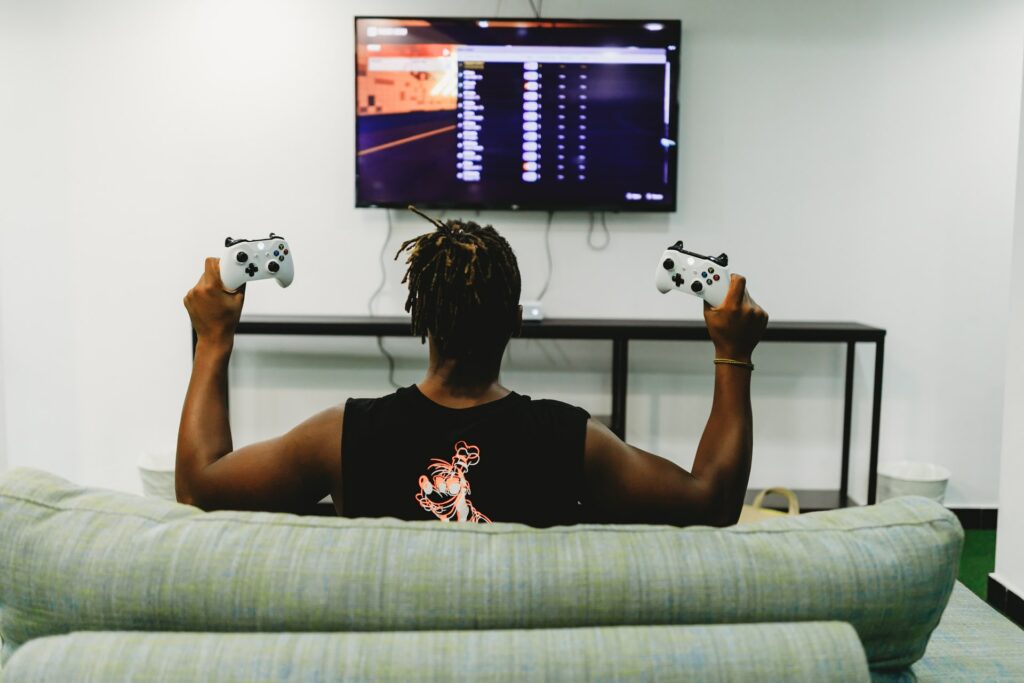 Someone sat on a sofa, pictured form behind, holding two Xbox controllers in front of a big screen tv, a setup that would be fun for online team building!