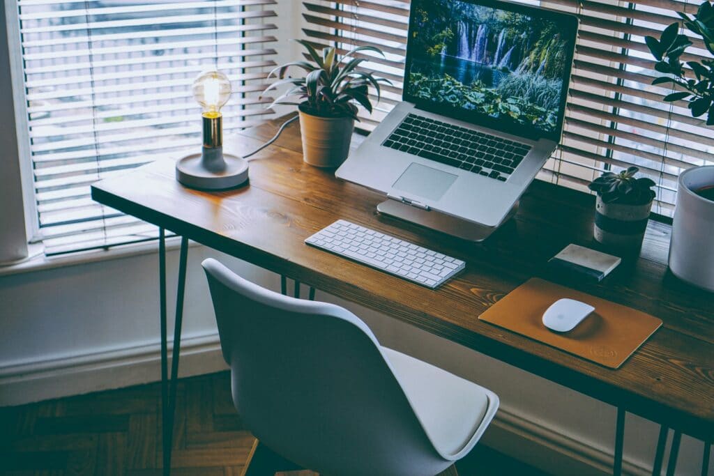 A productive workspace that includes a laptop on a riser, a light some potted plants and a keyboard on a simple desk with a comfortable chair.