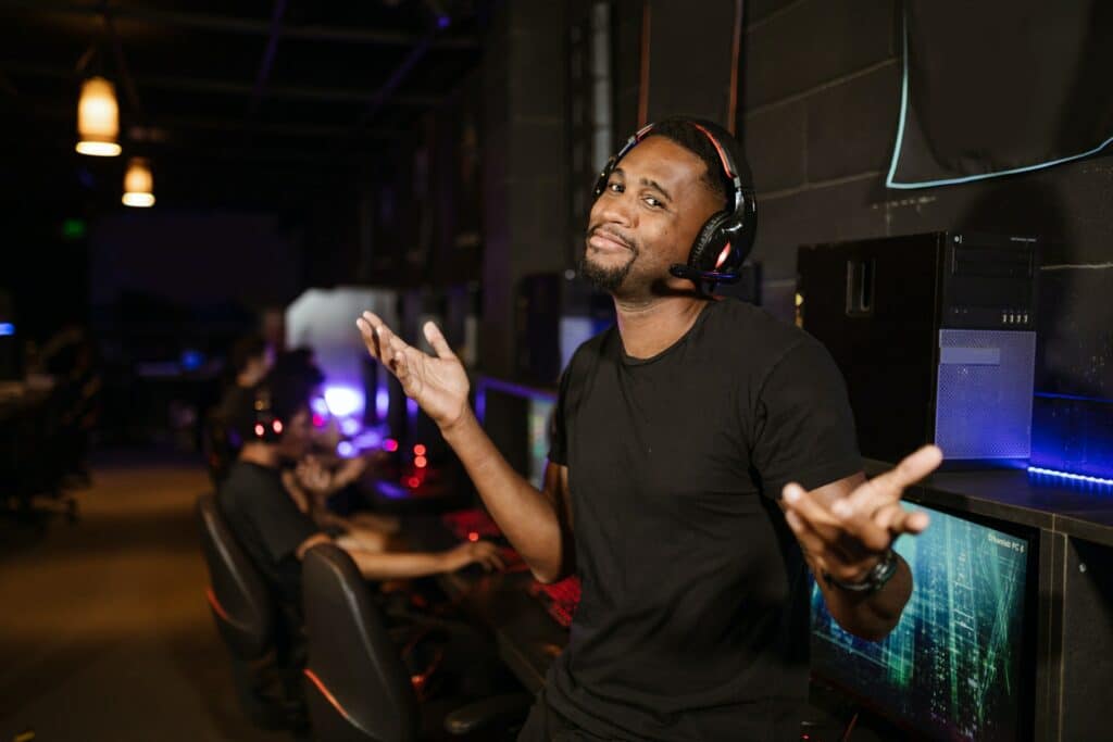 A person with a gaming headset on stood in front of a row of gaming PCs in a large room with two other people next to them, they are shrugging but smiling thinking about reusing content.