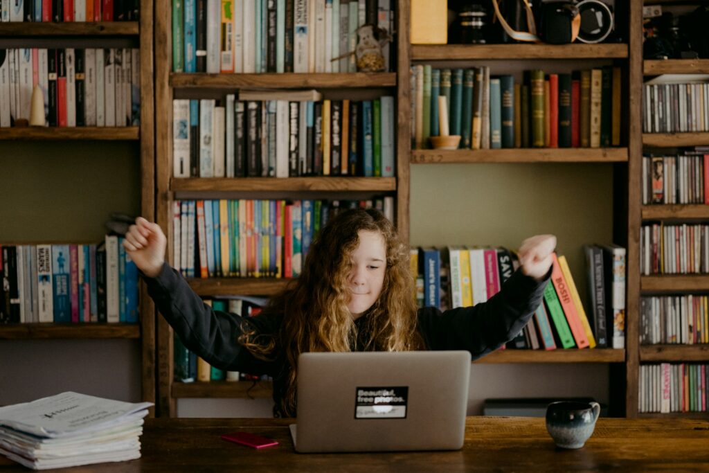 A child is sat facing a laptop with their arms outstretched with a bookcase full of books behind them. On the desk is a phone, a pile of papers and a mug as they take part in their secure online class.