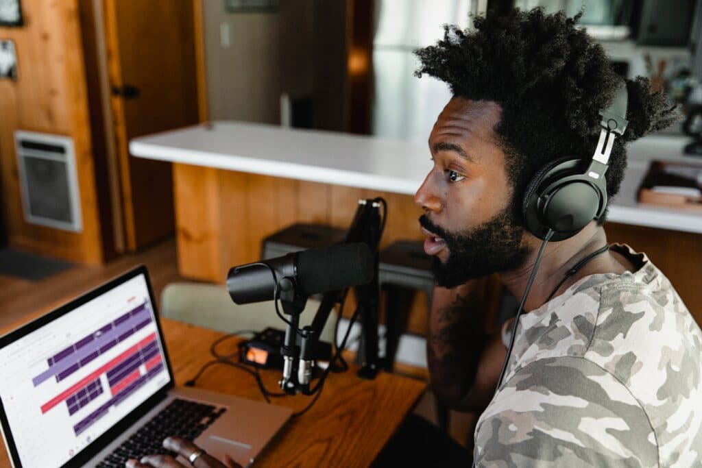 A content creator sat at a desk with headphones on talking in to a Shure microphone connected to a mic stand and a laptop for professional microphone sound.