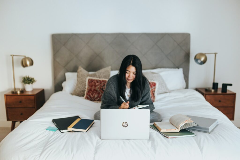 A woman sits on her bed with her notebooks spread out using a laptop in her bedroom, one of the benefits of working from home