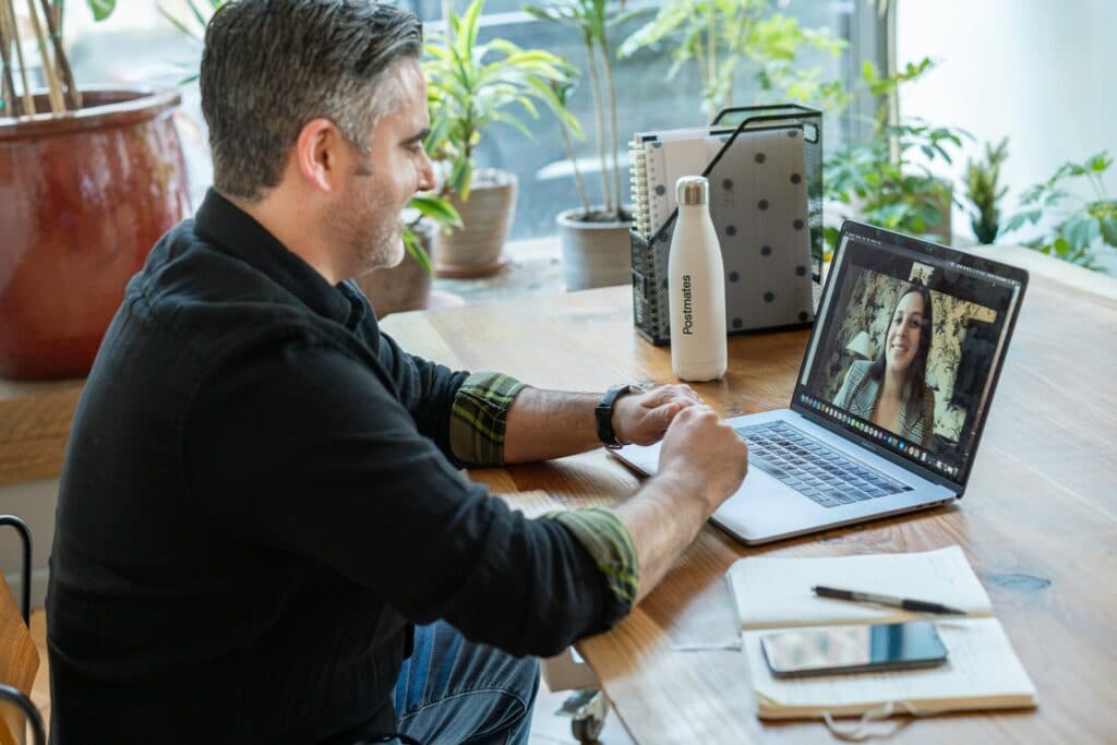 A person, well lit, is sat in front of a laptop on a call with someone, knowing the best lighting for a webcam. A notebook is on the table as well as a water bottle and some files.