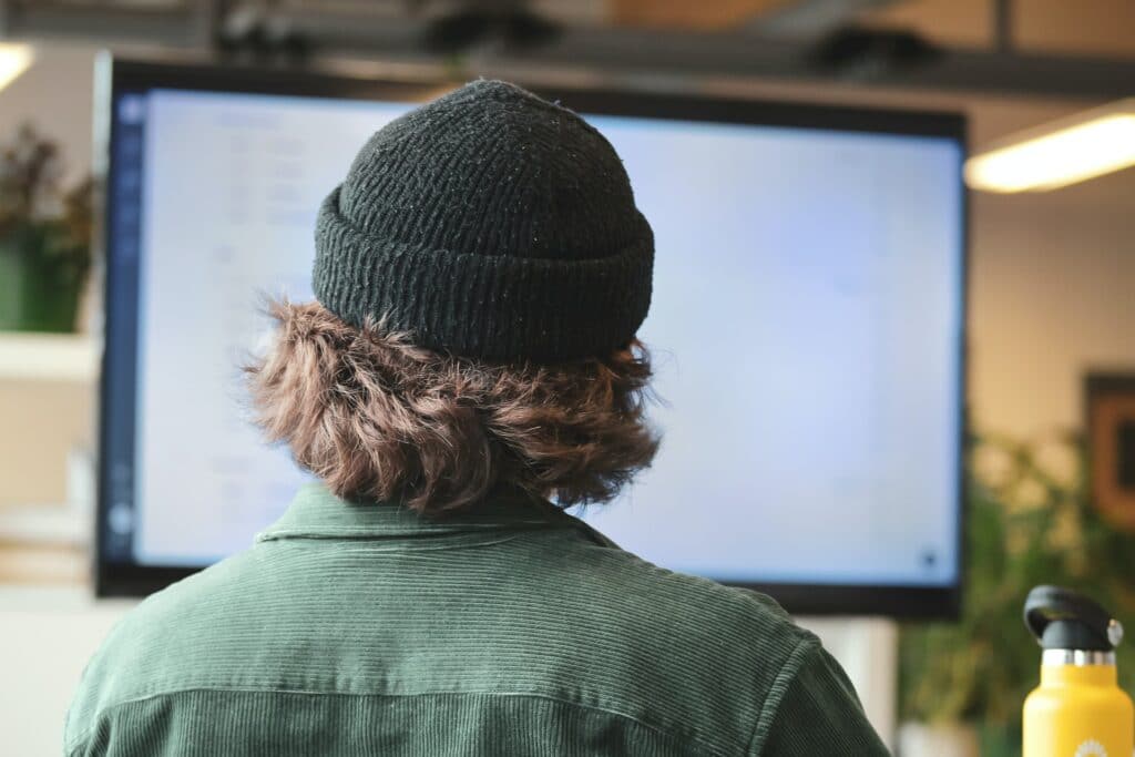 A person wearing a beanie with their back to the camera and a flask on their desk as they use the best work from home apps.