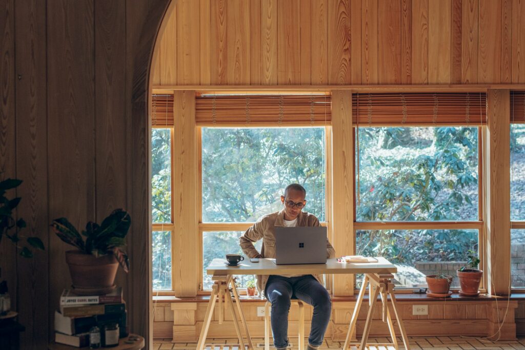 A person sits at a desk in thei spacious home with windows behind them showing trees. A laptop is on their desk as they use the best work from home apps.