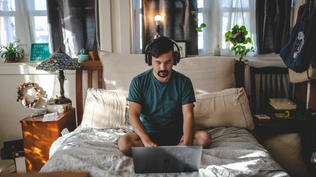A man working from home, sat on his bed cross legged with headphones on using his laptop as part of a hybrid office.