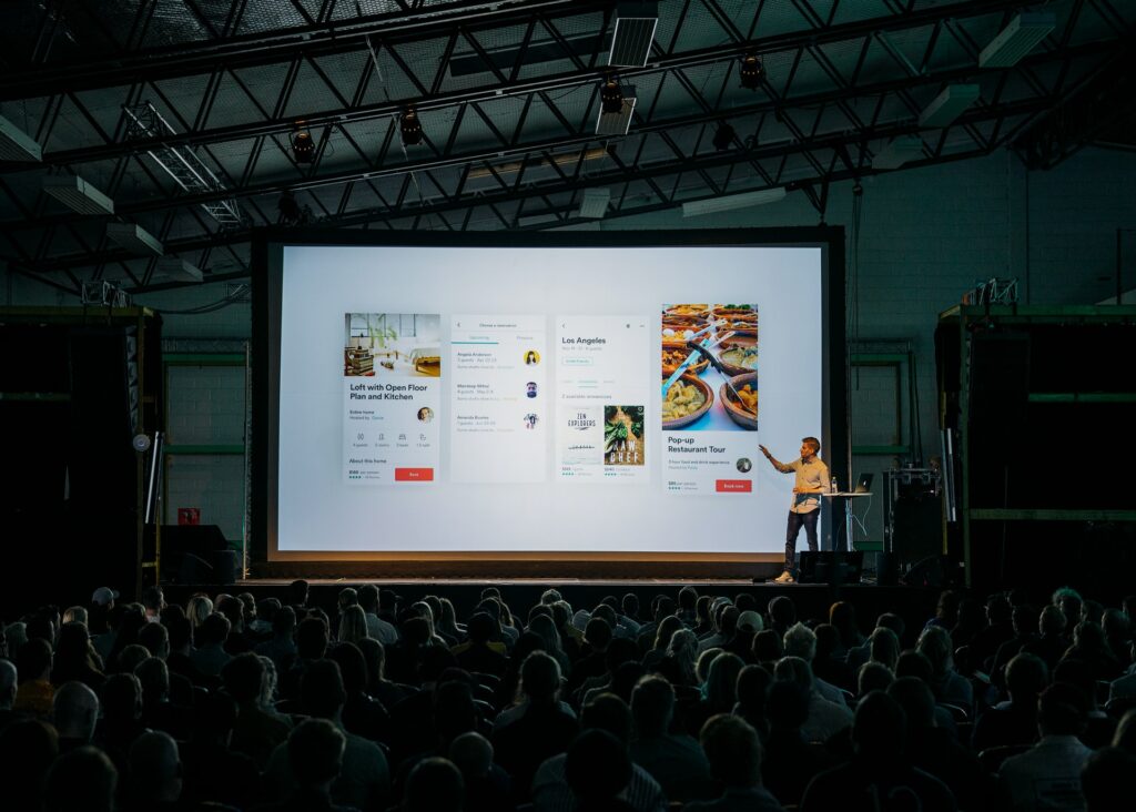 A person stood on the stage of a hybrid conference presenting a slide to a large watching audience on a big screen while also broadcasting to viewers watching at home online.
