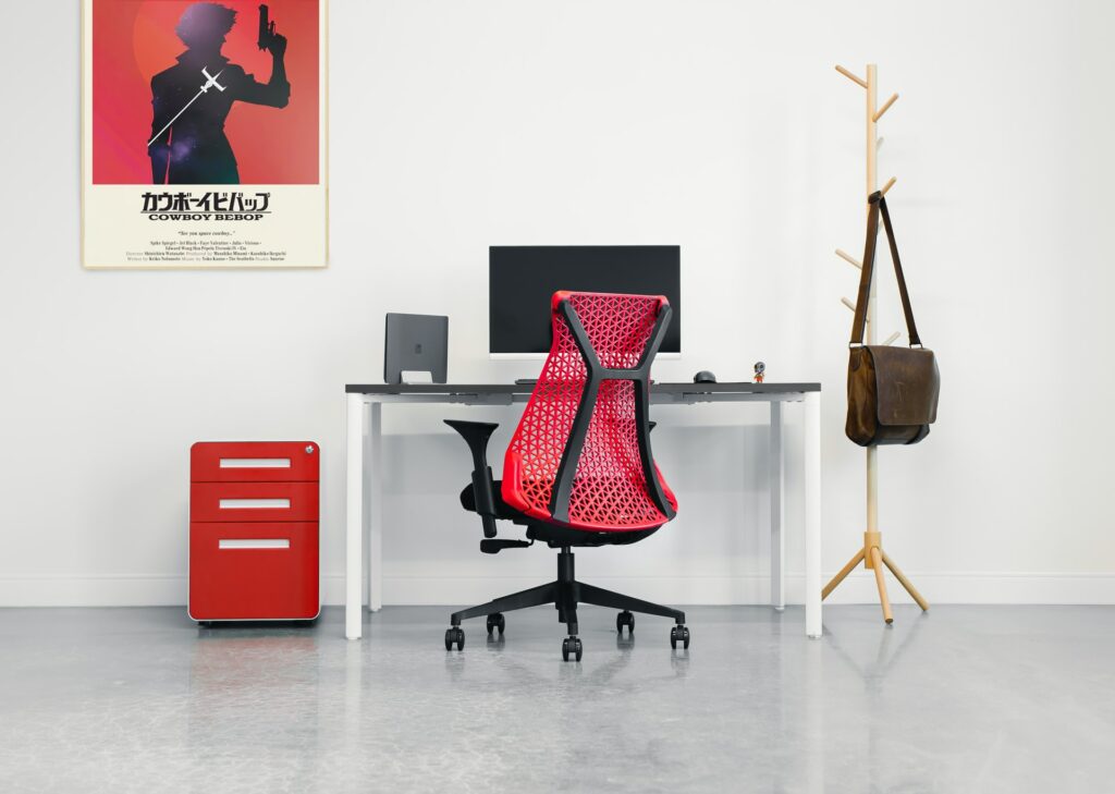 Office chair upgrades could include this red chair which is in front of a desk with a laptop connected to a monitor, a red filing cabinet is next to the desk and a large framed poster is on the wall.
