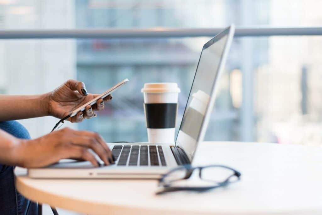 A person using a smartphone next to their laptop which is on the table in front of them next to a cup of coffee they could be using tools for remote workers on both!