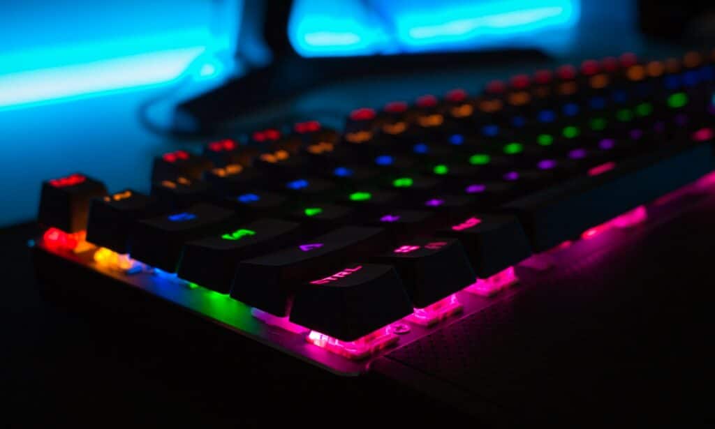 An affordable gaming keyboard close up with it's RGB lights on showing multiple colours, you can see a monitor stand in the background and a backlight.