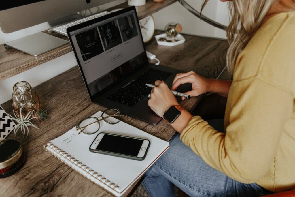 A person sat at their desk using a laptop computer with a notepad, phone and glasses next to them looking for work from home hacks.